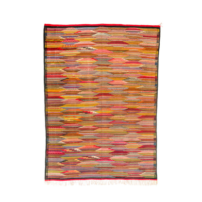 A rectangular woven moroccan rug from taznakht with a dense horizontal striped cubist pattern in warm tones of red, orange, yellow, beige, and brown, finished with a dark border and fringe.