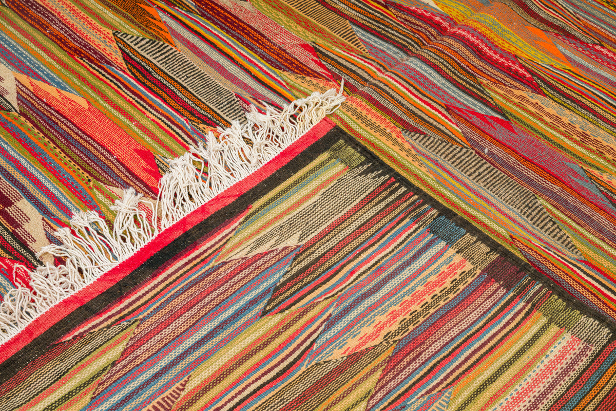 A close-up of a colorful woven  moroccan rug from taznakht with diagonal striped cubist patterns in red, orange, yellow, green, blue, and brown, featuring a white fringed edge.