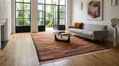 A large rectangular area rug from taznakht with a multicolor striped pattern in red, orange, yellow, green, and blue, styled in a modern living room with a gray sofa and wooden floors.