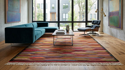 A large rectangular moroccan area rug from taznakht with a multicolor cubist abstract pattern in red, orange, yellow, green, and blue, styled in a modern living room with a teal sectional sofa, wooden coffee table, and large windows.