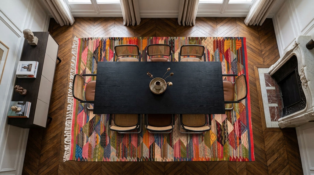 A top-down view of a large, vibrant multicolor handwoven moroccan rug from Taznakht  in a classic European dining room with herringbone wood floors, a black wooden table, and six chairs.