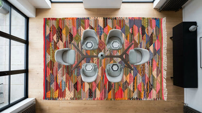 A top-down view of a large, vibrant handwoven moroccan rug from Taznakht in a modern room with light wood floors, surrounded by six gray chairs and a central table.