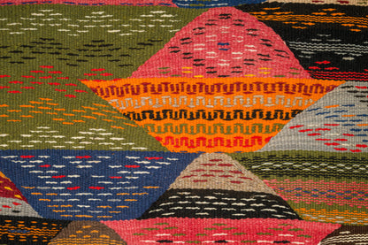 A close-up of a handwoven moroccan rug from Taznakht with a geometric patchwork design in multiple colors, including pink, orange, blue, green, black, and beige.