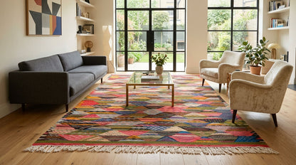A large, vibrant handwoven cubist moroccan rug from Taznakht  with multicolor patterns and white fringe, styled in a bright, modern living room with a gray sofa, beige armchairs, and a glass-top coffee table.