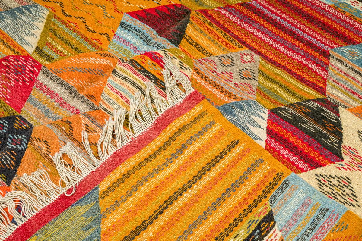 A close-up of a colorful woven moroccan rug from taznakht with geometric patchwork panels, fringe trim, and striped patterns in orange, yellow, red, blue, beige, and black.