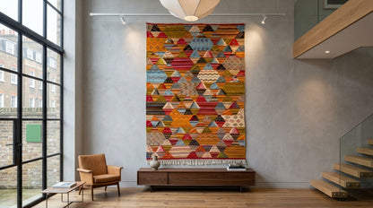 A large geometric wall hanging  using a handwoven moroccan rug with a colorful cubist pattern of triangles and diamonds, displayed in a modern loft-style room with a wooden floor, gray concrete walls, a brown armchair, and a staircase.