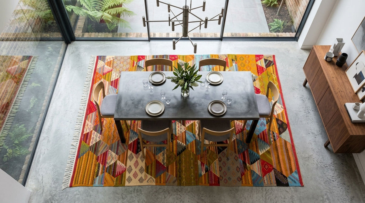 A colorful handwoven cubist area rug in a modern dining room setting, featuring a dark gray rectangular dining table and four wooden chairs on top, with a chandelier and large windows in the background.