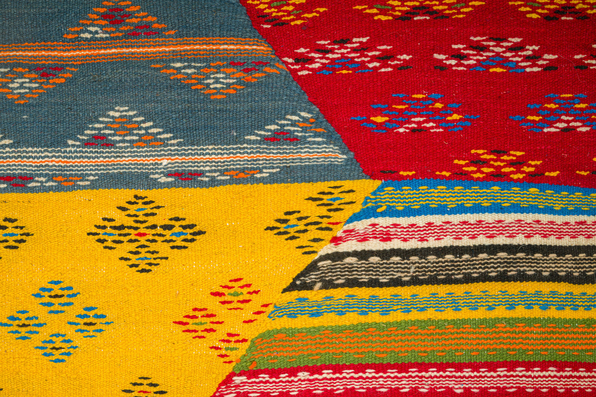 A close-up of a colorful woven moroccan rug from taznakht with geometric cubist patterns, showing sections of red, yellow, blue, and striped multicolor fabric.