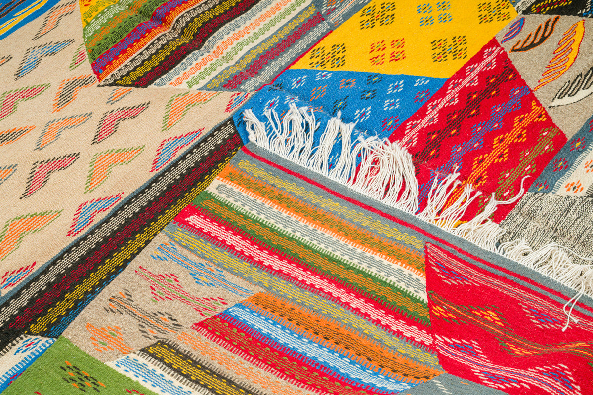 A close-up of a colorful handwoven textile or  moroccan rug with geometric and striped patterns in red, blue, orange, green, yellow, beige, and white, including visible fringe on one section.