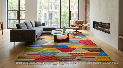 A colorful geometric moroccan rug with a multicolor abstract cubist pattern and fringe, styled in a modern living room with a gray sectional sofa, glass coffee table, and fireplace.