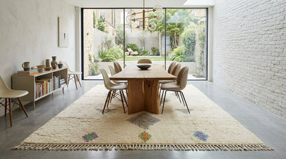 A large cream-colored area rug with colorful geometric patterns and fringe trim, styled in a modern dining room with a wooden table and chairs.