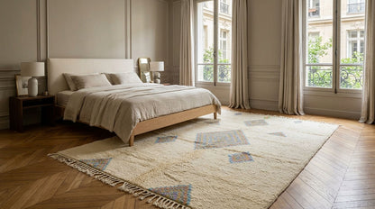 A large cream-colored area rug with a geometric diamond pattern in muted blue, gray, and beige tones, styled in a modern bedroom with a wooden bed and herringbone wood floors.