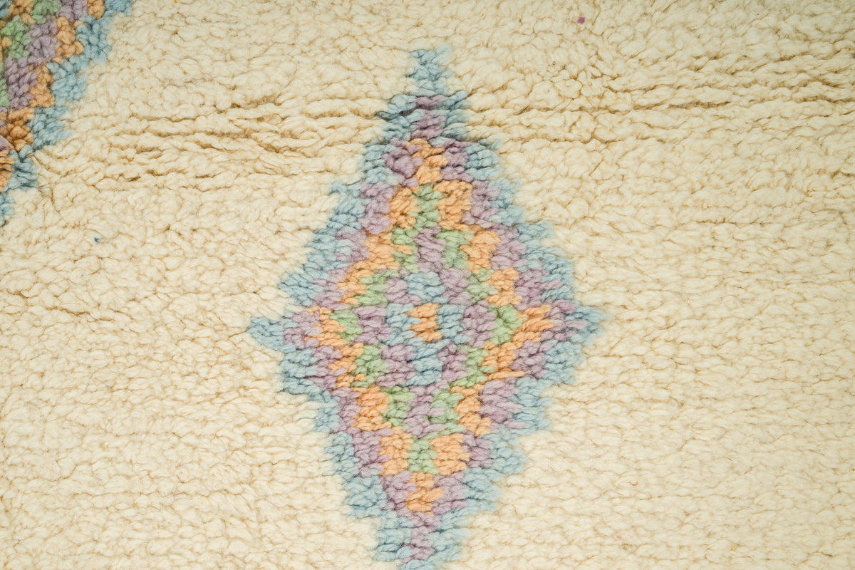 Close-up of a cream-colored rug featuring a central diamond-shaped geometric pattern in pastel shades of blue, purple, and orange.