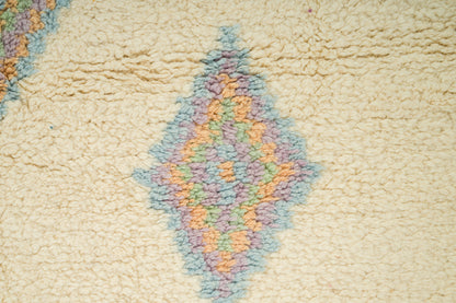 Close-up of a cream-colored rug featuring a central diamond-shaped geometric pattern in pastel shades of blue, purple, and orange.