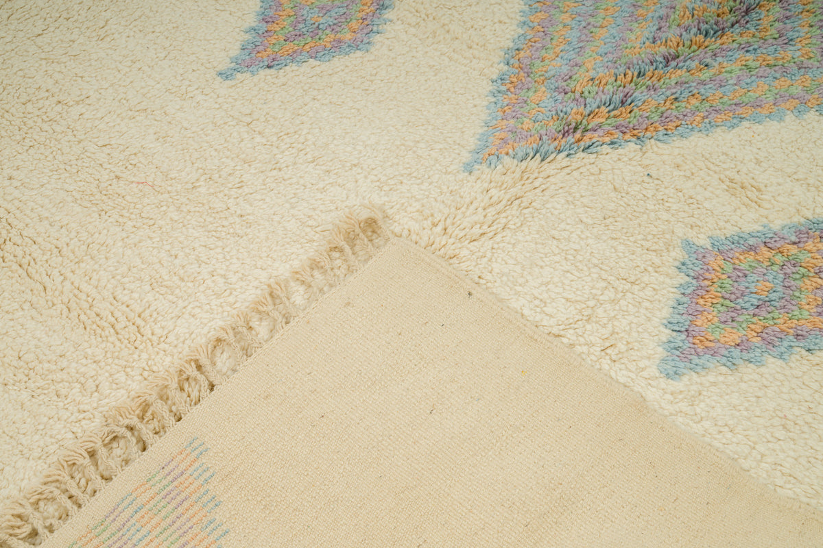 Close-up of a cream-colored shag rug with a textured geometric pattern in pastel blue, orange, and yellow, featuring a fringed edge and a woven backing.