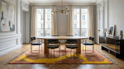 A modern dining room featuring a round wooden dining table surrounded by six black upholstered chairs with gold metal frames, set on a large abstract patterned area rug in warm tones. The room has classic white paneled walls, tall windows with beige curtains, and a chandelier.
