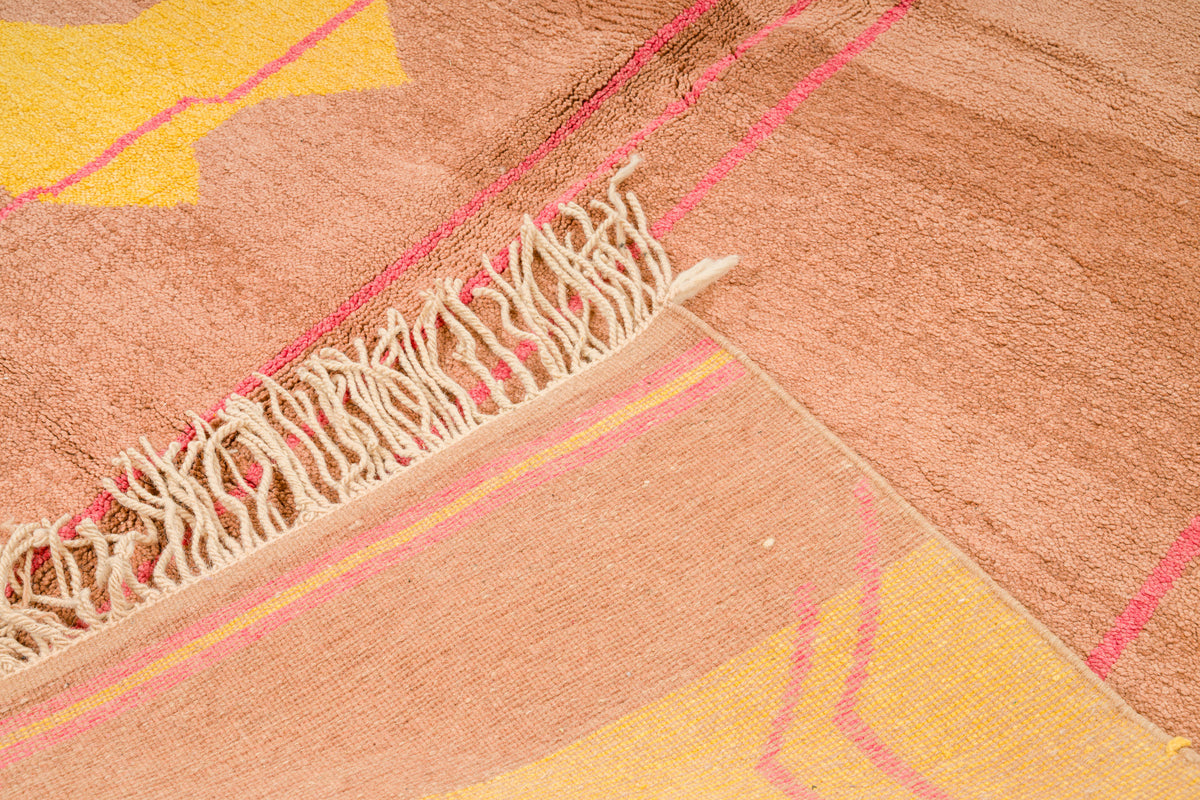 Close-up of a Moroccan rug with cream fringe, showing a warm tan and peach base with yellow geometric sections and thin pink lines.
