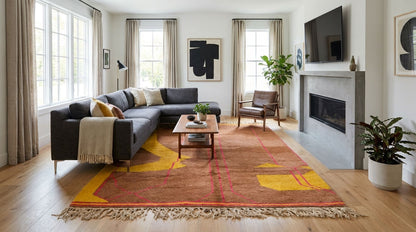 A large area rug with a red, orange, and yellow abstract pattern and fringed edges, styled in a modern living room with a dark gray sectional sofa, wooden coffee table, and light wood floors.