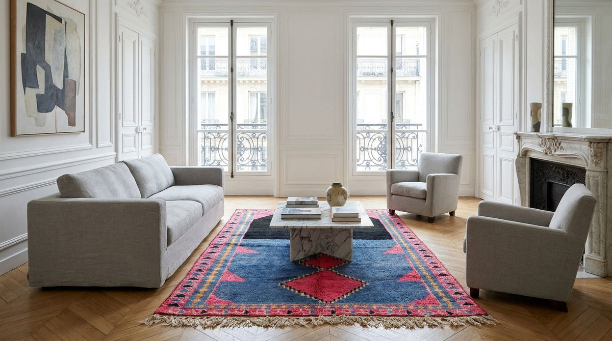 A bright, elegant living room featuring a light gray fabric sofa, two matching armchairs, a marble coffee table, and a prominent blue and red patterned oriental rug on a wooden floor.