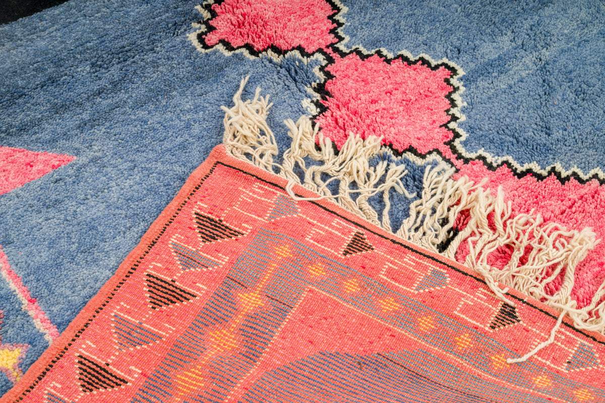 A close-up of a Azilal moroccan rug showing a blue field with bright pink geometric motifs outlined in black, meeting a coral-red patterned section with cream fringe.