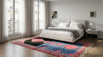 A modern bedroom featuring a large area rug with a blue, pink, and black geometric pattern on a herringbone wood floor. The room is styled with a light gray upholstered bed, white bedding, and matching gray nightstands and lamps.