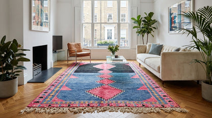 A large rectangular Moroccan Azilal area rug with a blue, pink, and red geometric tribal pattern and fringe, styled in a bright modern living room with a beige sofa, brown leather armchair, and potted plants.