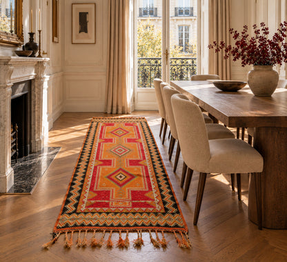 A long geometric patterned wool runner rug in red, orange, yellow, and black is styled on a wooden floor in a bright, elegant dining room with a wooden table, beige chairs, and a fireplace.