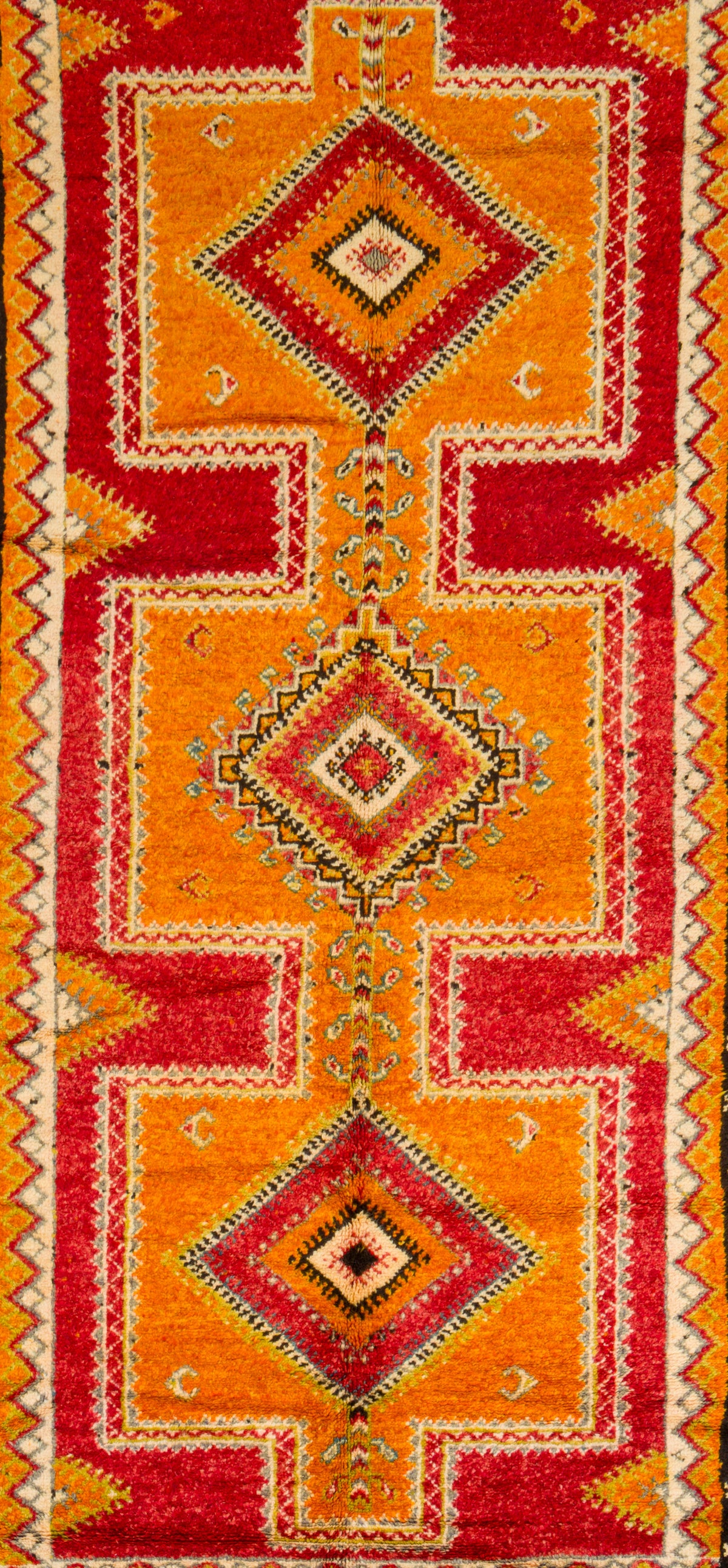 A close-up of a patterned textile or rug featuring three large stacked diamond motifs in red, orange, yellow, and cream, with geometric border details.
