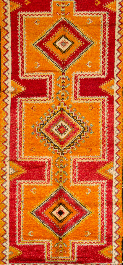 A close-up of a patterned textile or rug featuring three large stacked diamond motifs in red, orange, yellow, and cream, with geometric border details.