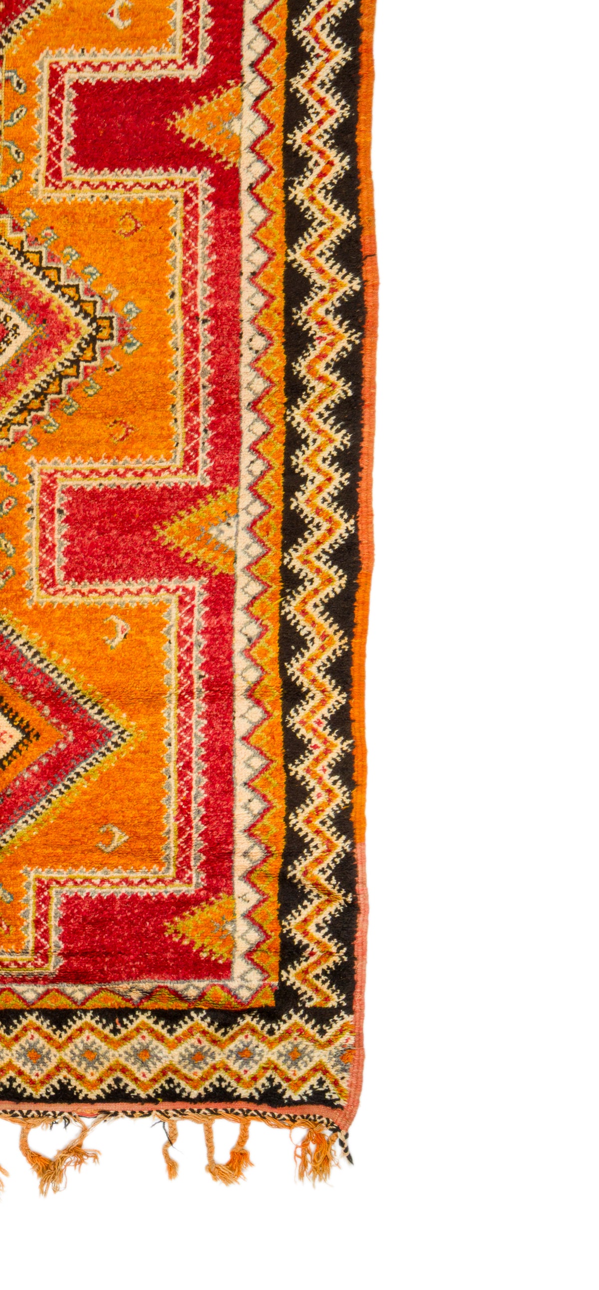 A close-up of a patterned textile or rug corner showing geometric stripes and borders in orange, red, black, and cream, with fringe tassels along the bottom edge, photographed against a white background.