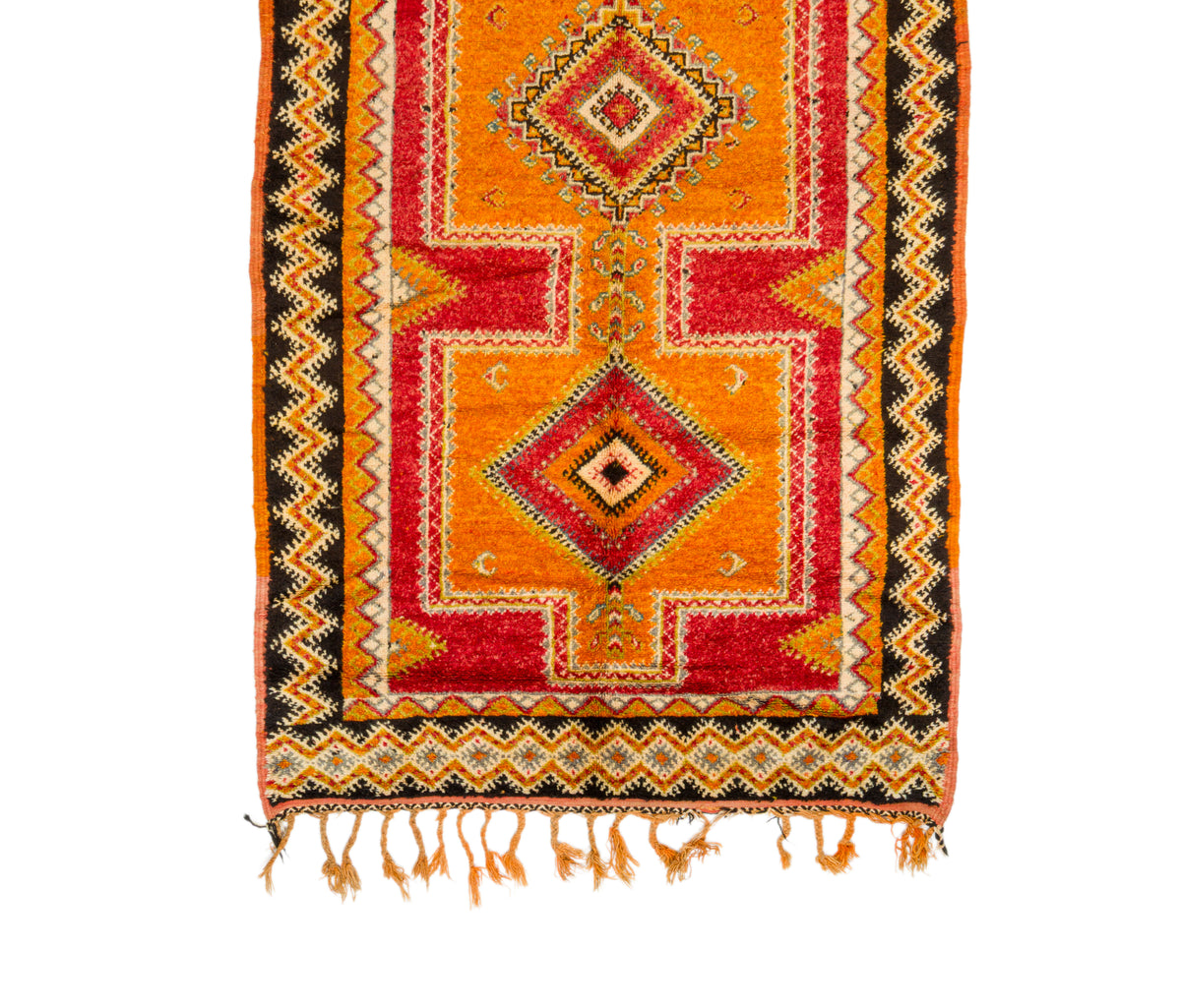 A close-up of a colorful Moroccan rug with geometric patterns, featuring orange, red, black, and beige colors, finished with a fringed hem.