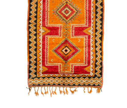 A close-up of a colorful Moroccan rug with geometric patterns, featuring orange, red, black, and beige colors, finished with a fringed hem.