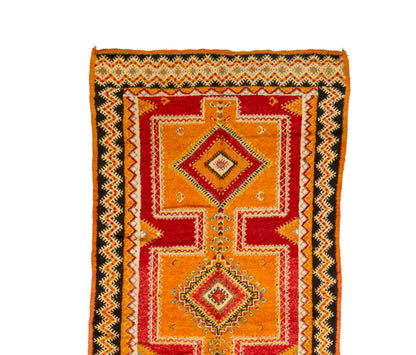 A vintage Moroccan wool rug with a geometric pattern in orange, red, black, and beige, shown flat against a white background.