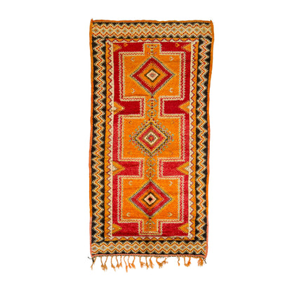 A rectangular woven Moroccan rug with a traditional geometric pattern in orange, red, yellow, and black, featuring three central diamond motifs and fringe tassels at the bottom, shown against a plain white background.