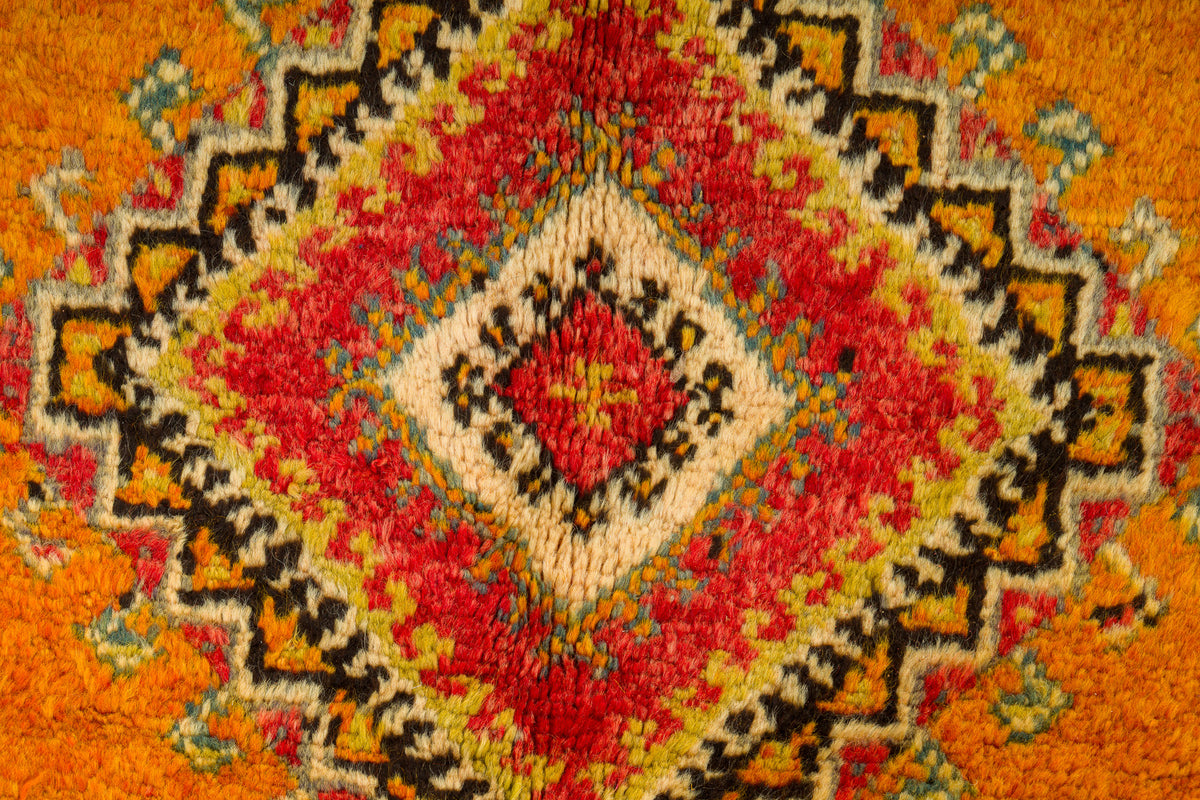 A close-up of a textured, hand-woven rug featuring a vibrant geometric diamond pattern in red, orange, beige, and black.