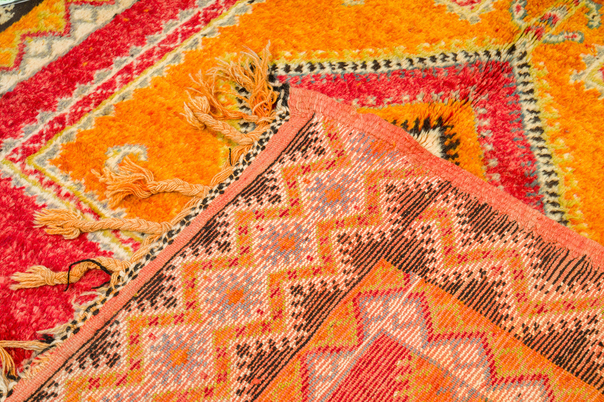 A close-up of a colorful patterned rug with geometric designs in orange, red, pink, brown, and cream, showing woven texture and tassels.