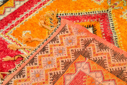 A close-up of a colorful patterned rug with geometric designs in orange, red, pink, brown, and cream, showing woven texture and tassels.