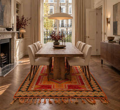 A rustic wooden dining table surrounded by cream upholstered chairs, set on a large red, orange, and black patterned area rug in a bright, elegant dining room with a fireplace and large windows.