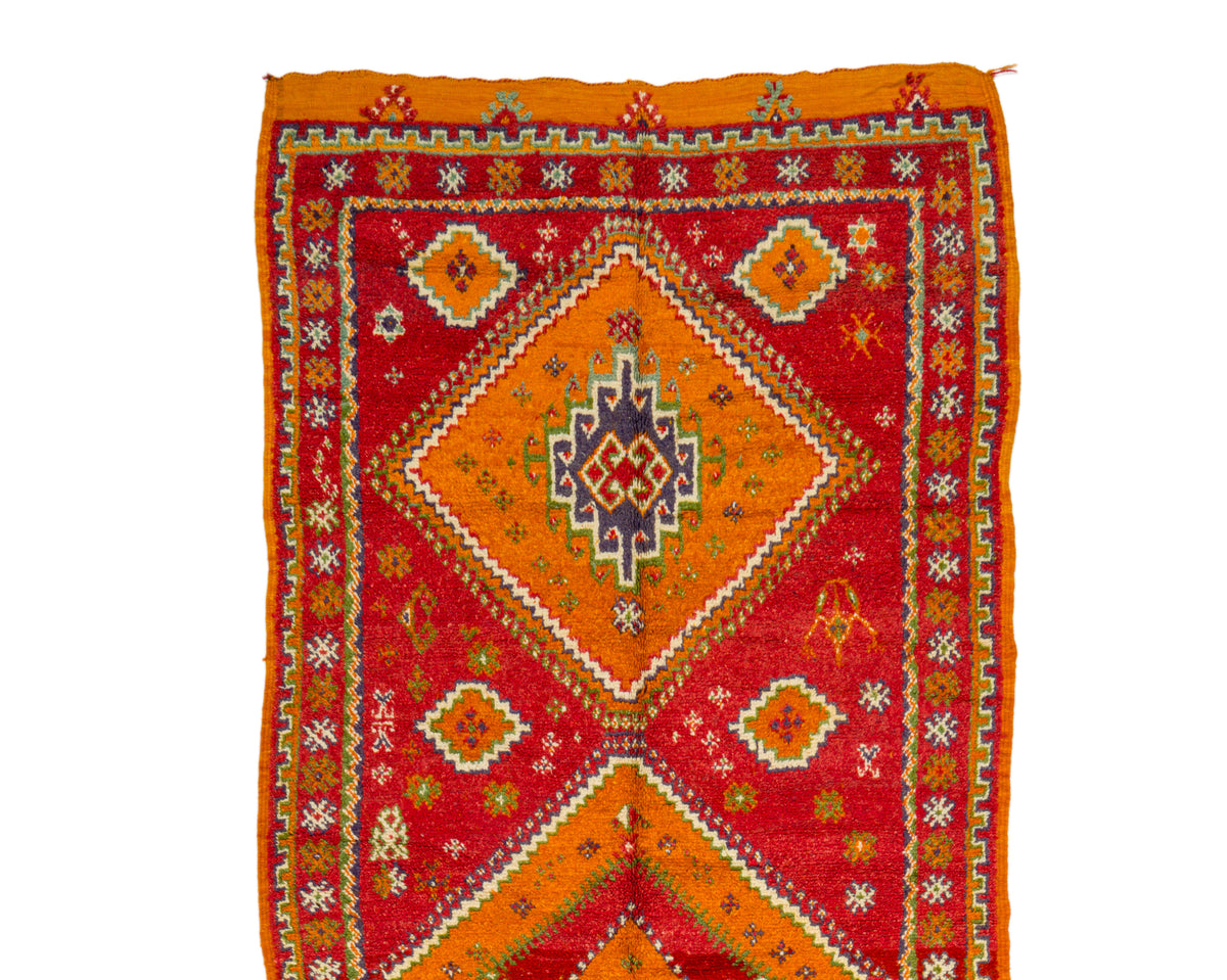 A square handwoven rug with a vibrant red and orange geometric pattern, featuring a central diamond motif and intricate border details in yellow, white, and blue, shown flat against a white background.