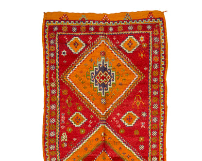 A square handwoven rug with a vibrant red and orange geometric pattern, featuring a central diamond motif and intricate border details in yellow, white, and blue, shown flat against a white background.