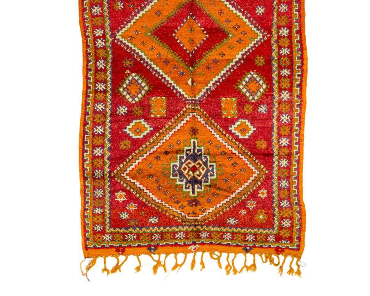 A rectangular woven rug with a bold geometric pattern in red, orange, yellow, and cream, featuring fringe tassels along the bottom edge, shown flat against a white background.