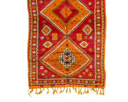A rectangular woven rug with a bold geometric pattern in red, orange, yellow, and cream, featuring fringe tassels along the bottom edge, shown flat against a white background.