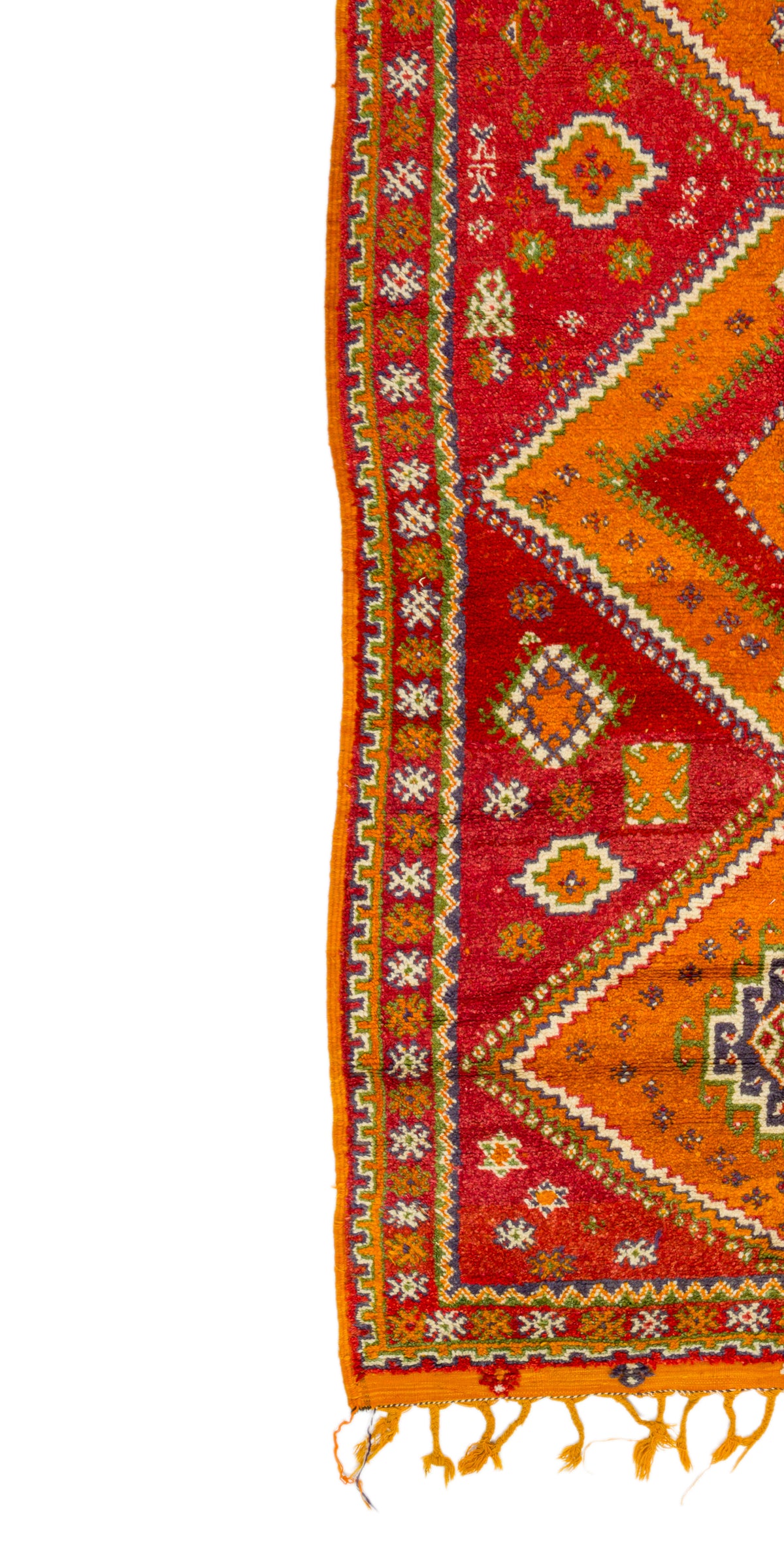 A close-up of a patterned Moroccan rug corner showing a red and orange geometric design with yellow, white, and blue accents and tassels along one edge, photographed against a white background.