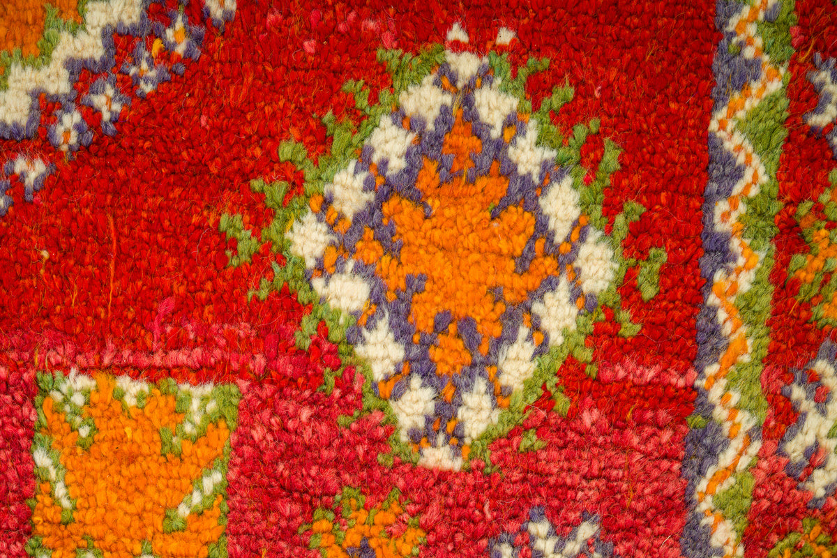 A close-up of a textured, loop-pile rug featuring a vibrant red background with a central medallion pattern in orange, cream, green, and purple.