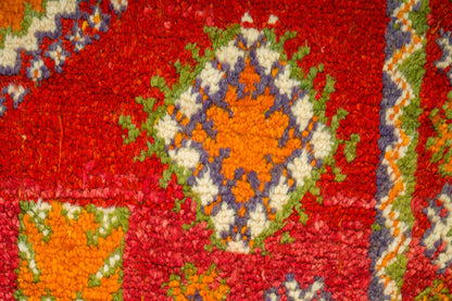 A close-up of a textured, loop-pile rug featuring a vibrant red background with a central medallion pattern in orange, cream, green, and purple.