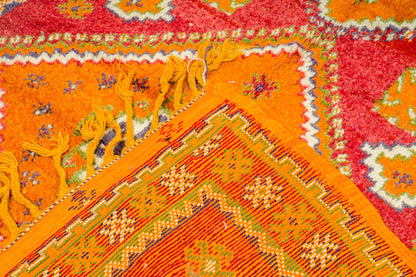 Close-up of a colorful patterned rug with bright orange, red, yellow, and cream geometric and floral motifs, including tassels along one edge.