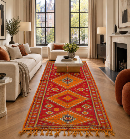 A long red and orange patterned runner rug with geometric diamond motifs and fringe, styled in a bright modern living room with a cream sofa, coffee table, and large windows.