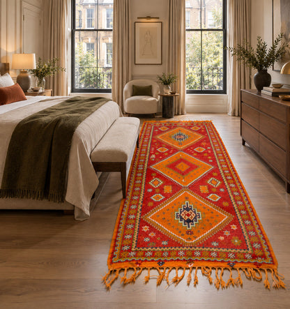 A long red and orange patterned runner rug with tassels is styled on a light wood floor in a bright, modern bedroom with a bed, dresser, and large windows.