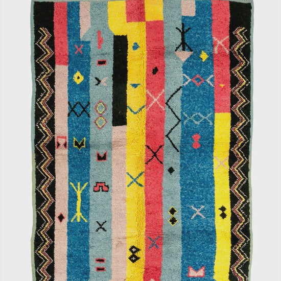 video of a rectangular hand knotted Moroccan rug with a multicolor geometric pattern featuring vertical stripes in blue, yellow, pink, black, and beige, finished with a fringed top and bottom edge.