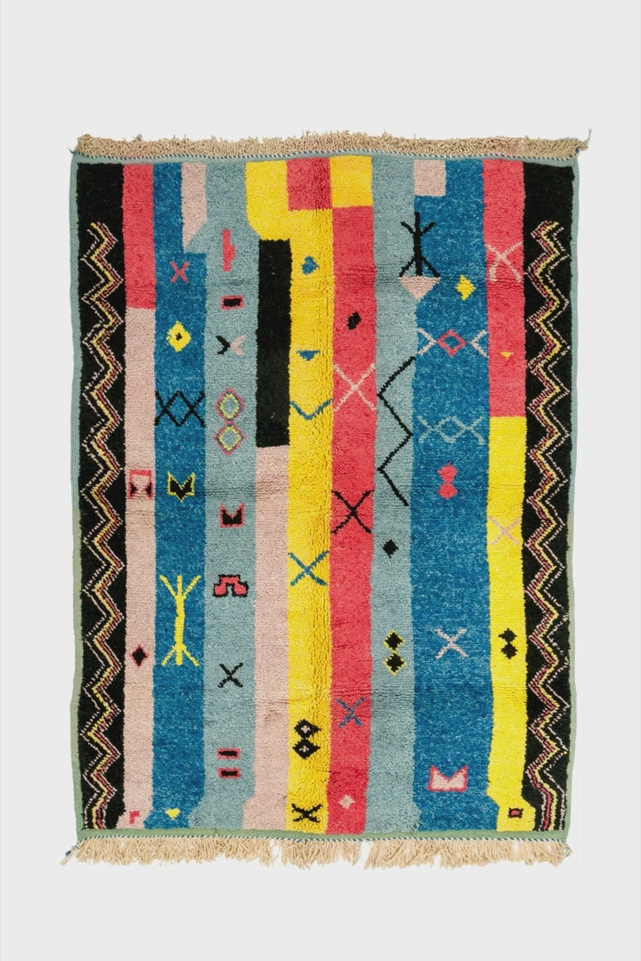video of a rectangular hand knotted Moroccan rug with a multicolor geometric pattern featuring vertical stripes in blue, yellow, pink, black, and beige, finished with a fringed top and bottom edge.